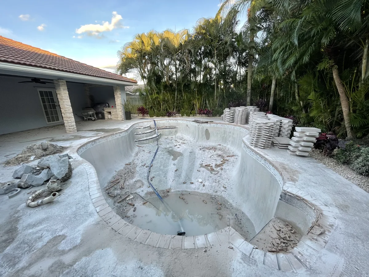 Freeform pool drained, with the old finish chipped out and pallets of new deck stone stacked behind it
