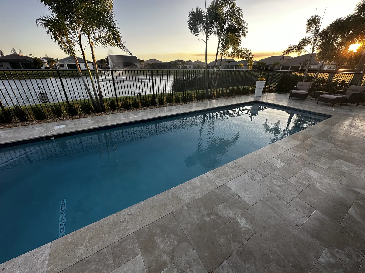 Rectangular pool with a wide silver travertine deck and loungers along a lake at sunset
